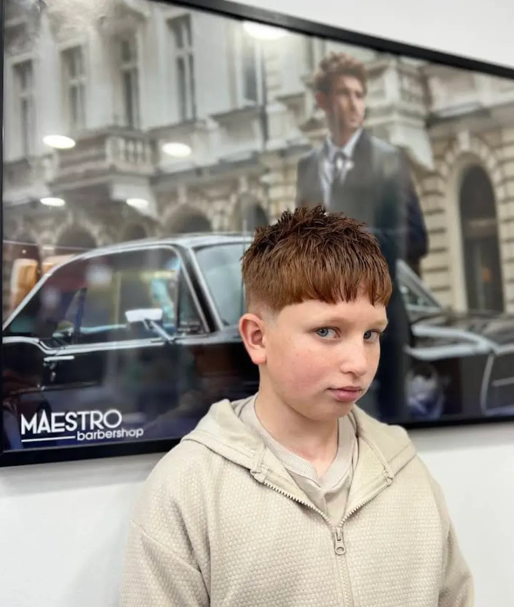 A boy with a fresh textured crop and mid fade, in front of the Maestro Barbershop wall print
